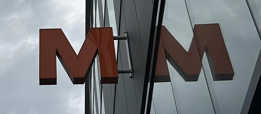Foto di una «M» arancione davanti a un negozio Migros, che si riflette sulla facciata vetrata. Anche il tempo uggioso si riflette sulla facciata. (Foto: area)
